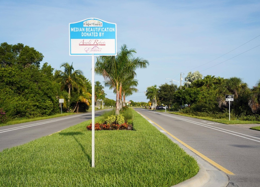 A street-level photo of a street median with decorative landscaping and a sign in the foreground with the words "median beautification donated by" and a script-style logo of a company below.